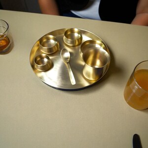 May include: A brass serving tray with a circular design, featuring multiple small bowls, a spoon, and a larger cup. Two glasses with liquids are on either side. The tray and items have a golden hue, set on a neutral-toned surface.