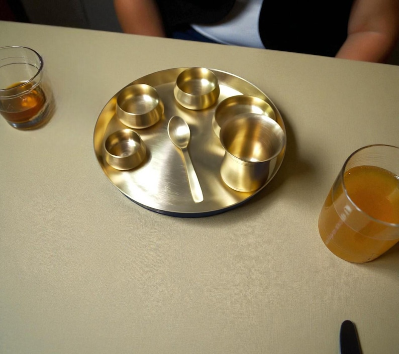 May include: A brass serving tray with a circular design, featuring multiple small bowls, a spoon, and a larger cup. Two glasses with liquids are on either side. The tray and items have a golden hue, set on a neutral-toned surface.