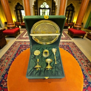 May include: A gold-coloured serving set presented in a green velvet-lined box. The set includes a round tray with floral designs, small bowls, and decorative pieces. The box is open, revealing the contents on an orange table.