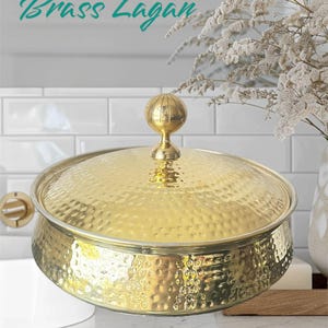 May include: A gold-colored brass Lagan bowl with a hammered texture and a clear glass lid. The lid is topped with a decorative spherical handle. The text "Brass Lagan" is written in teal script above the bowl.