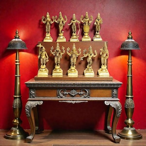 May include: A collection of gold-coloured deity statues displayed on a wooden table against a red wall. Two ornate brass lamps stand on either side. The statues are arranged on a wooden shelf, featuring detailed carvings and intricate designs.