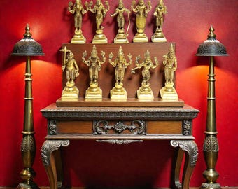 Op de afbeelding: Een display van goudkleurige godenbeelden op een houten tafel, tegen een rode muur. Twee sierlijke messing lampen flankeren de tafel. De beelden zijn gerangschikt op een houten plank, met gedetailleerde gravures en ingewikkelde ontwerpen.