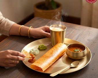 May include: A brass dinnerware set consisting of a large round plate, a small bowl, a tall tumbler, and a spoon. The plate holds a rolled crepe, three small dollops of chutney, and the bowl contains a reddish-brown liquid with green garnish.