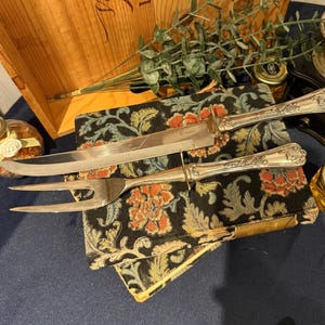 Puede incluir: Juego de cuchillo y tenedor de trinchar plateados con mangos ornamentados. La hoja del cuchillo es curva y el tenedor tiene dos púas. El juego descansa sobre una tela estampada floral. Una caja de madera y pequeños frascos están en el fondo.