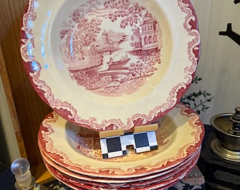 Pode incluir: Uma pilha de pratos antigos creme com um design vermelho. Os pratos apresentam uma paisagem cénica com edifícios e árvores, e uma borda vermelha decorativa. Os pratos são empilhados em cima de livros antigos.