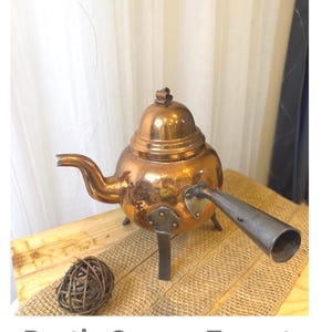 May include: A rustic copper teapot with a spout and handle. The teapot has a rounded body and sits on three metal legs. The image also includes a decorative woven ball and the text "Rustic Copper Teapot."