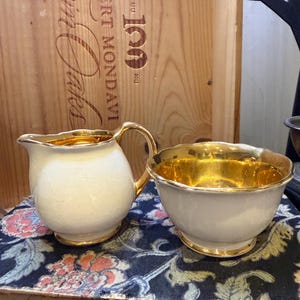 Könnte beinhalten: Ein weißes Sahne- und Zuckerdosen-Set mit goldenen Verzierungen. Der Sahnekännchen hat einen Griff und eine abgerundete Form. Die Zuckerdose hat einen gewellten Rand und eine goldene Innenseite. Das Set steht auf einem Buch mit floralem Muster.