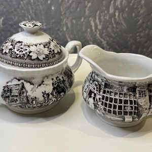 Puede incluir: Un juego vintage de azucarero y jarra de crema de cerámica blanca con un diseño floral negro y marrón. El azucarero tiene una tapa y ambas piezas presentan una escena de un edificio con un letrero que dice "Coach & Cri".