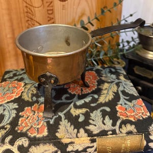 Op de afbeelding: Kleine koper- en zilverkleurige metalen pot met een zwarte handgreep en drie poten. De pot staat op een boek met een bloemenpatroon in tinten oranje, groen en blauw.