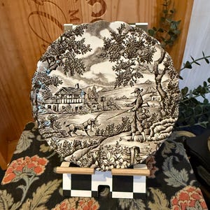 Può includere: Un piatto decorativo in bianco e nero con una scena pastorale con una casa, alberi e figure. Il piatto ha un bordo smerlato ed è esposto su un supporto di legno. Il piatto è ambientato su uno sfondo a motivi floreali.