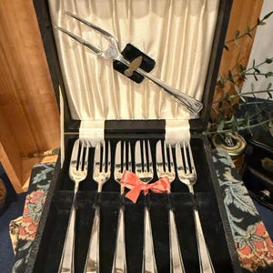 Pode incluir: Um conjunto de talheres vintage em prata, numa caixa preta com forro de cetim creme. O conjunto inclui vários garfos e um garfo de servir, todos com acabamento prateado brilhante. Um pequeno laço coral adorna os garfos.