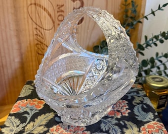May include: A clear, cut-glass basket with a handle. The basket features intricate geometric patterns and a starburst design on the base. The handle is also detailed with a textured pattern. The basket is sitting on a floral patterned surface.