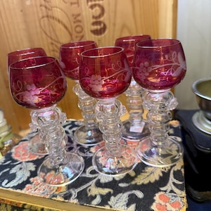 May include: A set of six ruby red wine glasses with clear, ornate stems. Each glass has a grape vine design etched into the bowl. The glasses are displayed on a floral patterned book.