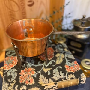 Puede incluir: Una pequeña cacerola de color cobre con un mango largo y un soporte de metal oscuro. La cacerola se encuentra sobre una pila de libros con un estampado floral en rojo, naranja y verde sobre un fondo negro. Se ve un tarro pequeño en el fondo.
