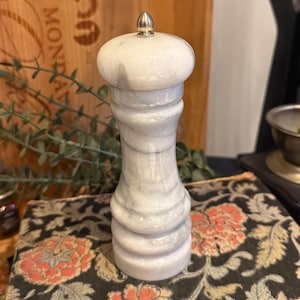 Può includere: Un macina sale o pepe in marmo con una parte superiore color argento. Il macinino è bianco con venature grigie e ha una parte superiore bulbosa e un corpo scanalato. Il macinino è appoggiato su una superficie a motivi floreali.
