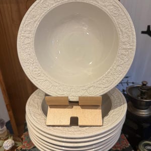 Könnte beinhalten: Ein Stapel weißer Keramikplatten und -schalen mit detaillierten floralen und scrollartigen Bordüren. Die Schalen sind tief und die Teller sind ordentlich gestapelt. Das Design ist klassisch und elegant, geeignet zum Servieren von Mahlzeiten.