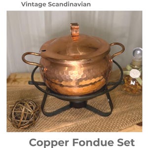 May include: Vintage copper fondue set with a hammered finish. The set includes a copper pot with a lid and handles, a black metal stand, and a small handle on the lid. The text "Vintage Scandinavian" and "Copper Fondue Set" are visible.