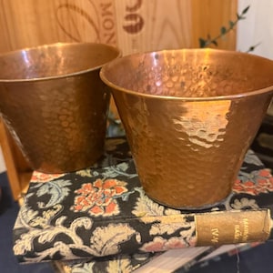 Puede incluir: Dos macetas de metal color cobre con textura martillada. Las macetas están sobre una pila de libros con estampados florales. Las macetas tienen forma acampanada y están vacías.