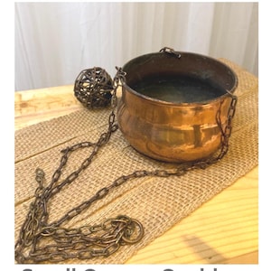 Puede incluir: Un pequeño caldero de cobre con una cadena, exhibido sobre una tela de arpillera sobre una superficie de madera. El caldero tiene forma redonda y un interior oscuro. El texto "Rustic Farmhouse Decor" y "Small Copper Cauldron" son visibles.