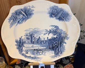 Op de afbeelding: Een witte keramische plaat met geschulpte rand en blauw transferontwerp. De plaat toont een gedetailleerde landschapsscène met een kasteel, bomen en een paardenkoets. Het ontwerp doet denken aan klassiek Engels aardewerk.