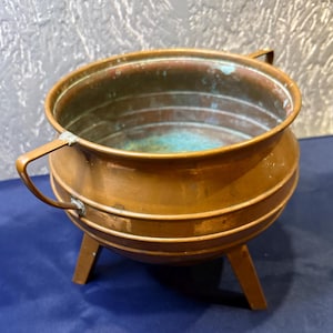May include: A copper pot with three legs and two handles. The pot has a worn, aged look and is perfect for a rustic or farmhouse decor.