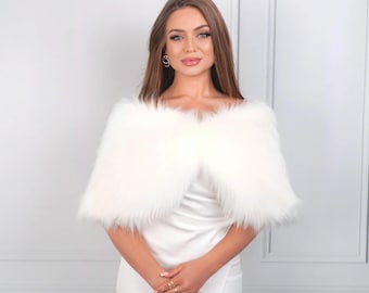 May include: A woman wearing a white faux fur shawl over a white dress. The shawl is off-the-shoulder and has a fluffy texture. The woman has long brown hair and is wearing gold jewelry.