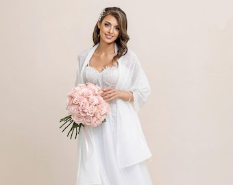 May include: A bride wearing a white wedding dress and a white shawl holds a bouquet of light pink roses.