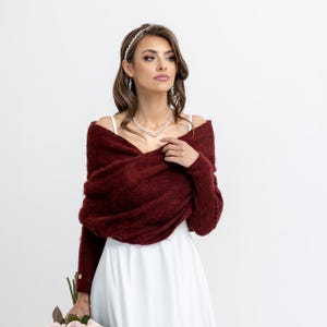 Pode incluir: Um xale de malha bordô com mangas compridas, usado sobre um vestido branco. O xale está drapeado sobre os ombros e amarrado na frente. A modelo está usando um colar de pérolas e uma faixa de cabeça de pérolas.