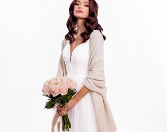 May include: A white sleeveless gown with a V-neckline and a full-length skirt, paired with a beige knit shawl draped over the shoulders. The garment is held alongside a bouquet of light pink roses with green stems and a light pink ribbon.