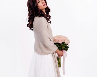 May include: A beige knit shawl draped over the shoulders, paired with a white skirt and a bouquet of light pink roses tied with a matching pink ribbon. The shawl features a soft, textured knit fabric with long, straight edges.