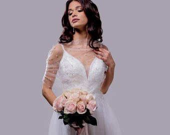 Puede incluir: Un vestido de novia blanco con escote pronunciado y mangas transparentes adornadas con perlas. La novia sostiene un ramo de rosas rosa claro atado con una cinta rosa. El vestido presenta detalles de encaje.