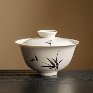 May include: White ceramic gaiwan tea set with a lid, featuring a black bamboo design. The tea bowl has a wide rim and a rounded base. The lid has a small handle. The set is on a dark surface.
