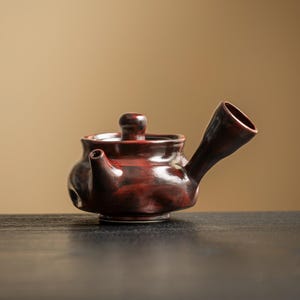 May include: A small, dark red ceramic teapot with a curved handle and a small spout. The teapot has a lid with a small knob and sits on a dark surface. The teapot is likely used for tea.