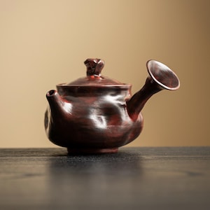 May include: A handcrafted, dark red ceramic teapot with a unique design. The teapot features a rounded body, a small spout, and a distinctive, flared spout. The lid is topped with a textured knob. The teapot is set against a neutral background.