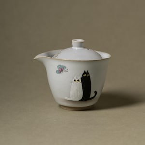 May include: A white ceramic tea bowl with a matching lid, featuring a painted illustration of a small white cat sitting next to a larger black cat. A small blue and pink butterfly is depicted above the cats on the side of the bowl.