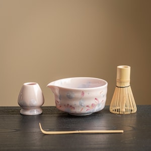 May include: A matcha tea set comprising a pink ceramic bowl with floral patterns, a small pink ceramic tea cup, a bamboo whisk, and a bamboo scoop. The set is displayed on a dark surface against a neutral backdrop.
