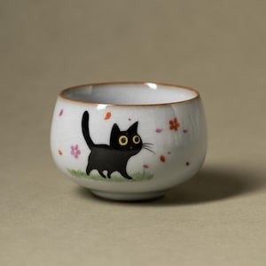 Può includere: Una tazza da tè in ceramica bianca con una finitura smaltata crepata e un'illustrazione dipinta di un gatto nero con grandi occhi gialli. Il gatto è raffigurato mentre cammina su un lembo di erba verde, circondato da piccoli petali di fiori rosa e arancioni.