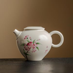 Peut inclure: Une théière en céramique blanche avec un couvercle et une poignée incurvée. La théière est décorée d'un motif floral avec des fleurs roses et des feuilles vertes. La théière est posée sur une surface sombre.
