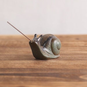 May include: A ceramic snail incense holder with a brown and green glaze. The snail has a hole in the top for an incense stick to be inserted.