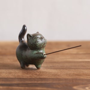 May include: A green and black ceramic cat incense holder with a stick of incense inserted into the holder.