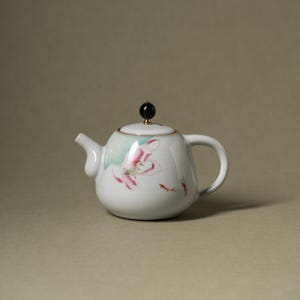 Op de afbeelding: Een witte keramische theepot met een handgeschilderd ontwerp van een roze lotusbloem en twee kleine oranje vissen. De theepot heeft een gebogen handvat, een korte tuit en een deksel met een kleine, ronde, zwarte knop op een gouden voet.