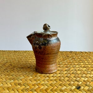 May include: A small, brown and black ceramic teapot with a lid. The teapot has a textured surface and a unique, rounded shape.