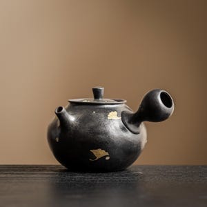 Puede incluir: Una tetera de cerámica negra mate con asa lateral y tapa a juego. La superficie presenta detalles de pan de oro dispersos en forma de hojas de ginkgo. La tetera tiene un cuerpo redondeado, un pico corto y un acabado oscuro y texturizado.