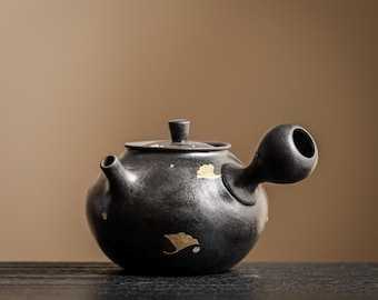 May include: A matte black ceramic teapot with a side handle and a matching lid. The surface features scattered gold leaf accents in the shape of ginkgo leaves. The teapot has a rounded body, a short spout, and a smooth, dark, textured finish.