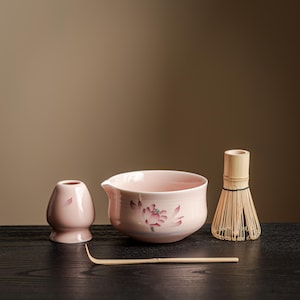 Op de afbeelding: Een matcha-theeset met een roze keramische kom met een bloemenontwerp, een bamboe klopper, een kleine roze keramische container en een bamboe lepel. De set is geplaatst op een donker houten oppervlak tegen een neutrale achtergrond.