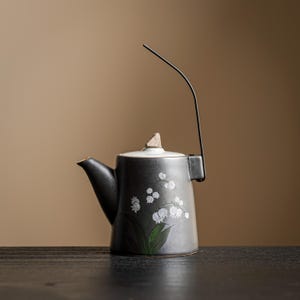 May include: A dark gray ceramic teapot featuring a hand-painted white lily of the valley floral design with green leaves. The teapot has a white lid with a small triangular knob and a tall, thin, black metal handle that extends upward.