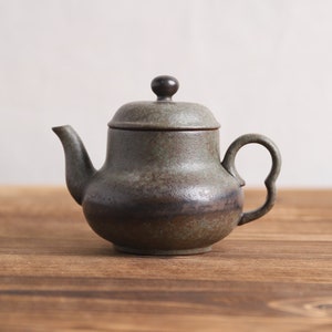 May include: A small, dark green teapot with a black lid and a black knob on top. The teapot has a slightly rounded body and a spout on one side. The teapot is sitting on a wooden surface.