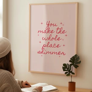 Peut inclure: Une affiche rose encadrée avec l'inscription "You make the whole place shimmer" en script rouge. Une petite plante en pot est posée sur un bureau en bois. Une personne lit un livre.