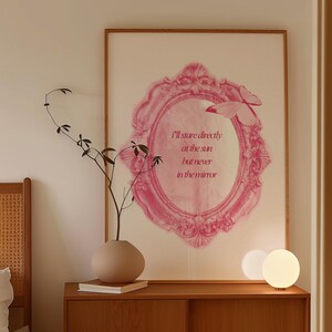 Pode incluir: Um interior de quarto com uma mesa de cabeceira de madeira, uma l&acirc;mpada branca, um vaso com um galho seco e uma impress&atilde;o emoldurada com uma moldura rosa adornada e o texto "Vou olhar diretamente para o sol, mas nunca para o espelho".