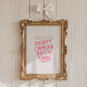 Pode incluir: Uma impressão emoldurada em dourado com uma fita branca pendurada no topo. A impressão apresenta o texto "There I'm a real tough kid, I am hustle or fall. They said, "Take you gotta fake it 'til you make it." and I did" em texto preto. O texto "LIGHTS CAMERA BITCH SMILE" está em texto rosa.